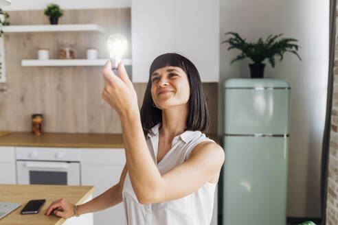 Smiling woman holding illuminated light bulb in kitchen at home - MEUF07203