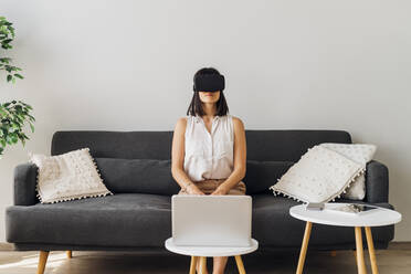 Woman using virtual reality glasses sitting on sofa at home - MEUF07223