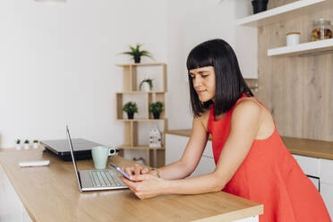 Freelancer woman using smart phone and laptop standing in kitchen - MEUF07234