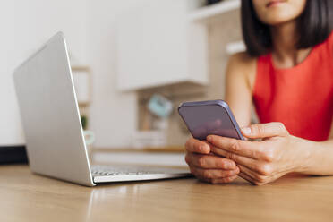 Hands of woman using smart phone with laptop at table at home - MEUF07236