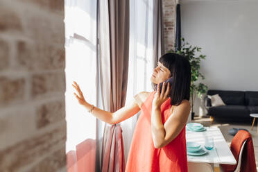 Woman talking on smart phone standing by curtain in living room - MEUF07239