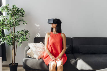 Frau mit Virtual-Reality-Brille sitzt zu Hause auf dem Sofa - MEUF07258