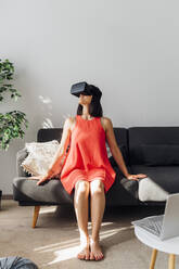 Frau mit Virtual-Reality-Simulator sitzt auf der Couch im Wohnzimmer - MEUF07259