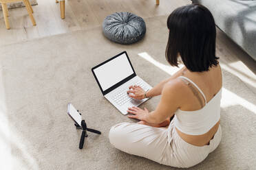Freiberufler, der einen Laptop mit einem Smartphone benutzt und zu Hause auf einem Teppich sitzt - MEUF07278
