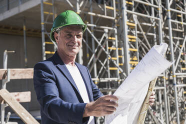 Smiling architect with blueprint standing at construction site - UUF26832