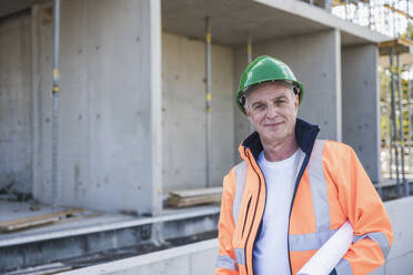 Smiling building contractor with blueprint standing at construction site - UUF26847