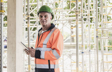 Senior contractor holding tablet PC standing in front of scaffolding - UUF26853