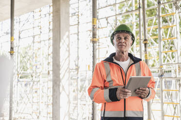 Thoughtful contractor holding tablet PC standing in front of scaffolding - UUF26855