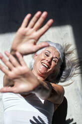 Happy mature woman with hands raised lying on floor - VEGF05695