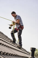 Handwerker mit Elektrowerkzeug und Seil auf dem Dach stehend - EBBF05680
