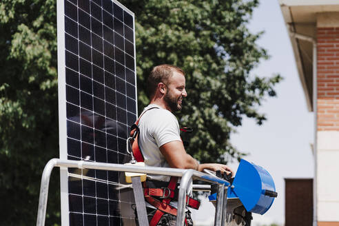 Smiling electrician with solar panel standing in hydraulic platform - EBBF05694