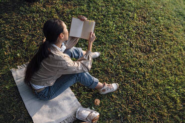 Woman reading book with food sitting on grass at park - VPIF06737