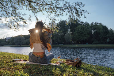 Woman sitting on grass with headphones by lake at park - VPIF06751