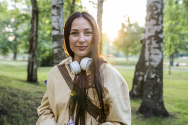 Smiling woman with headphones standing in park - VPIF06787