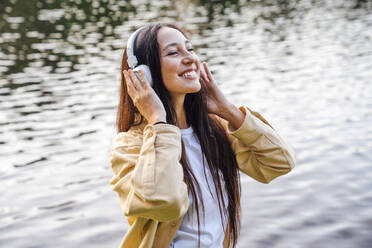 Smiling woman enjoying music with headphones in front of lake - VPIF06789