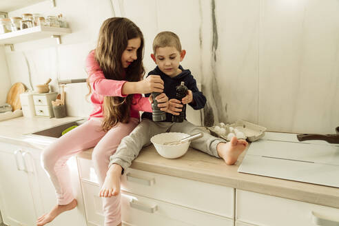 Mädchen mit Bruder hält Shaker über Schüssel auf Küchentisch zu Hause sitzen - OSF00396