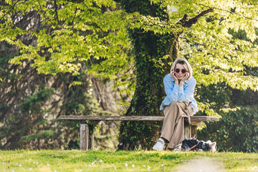 Reife Frau mit Sonnenbrille sitzt mit dem Kopf in den Händen auf einer Bank im Park - OMIF00967