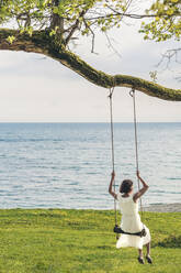 Woman sitting on swing by sea - OMIF00968