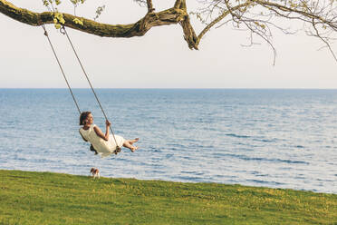 Woman swinging over grass by sea - OMIF00969