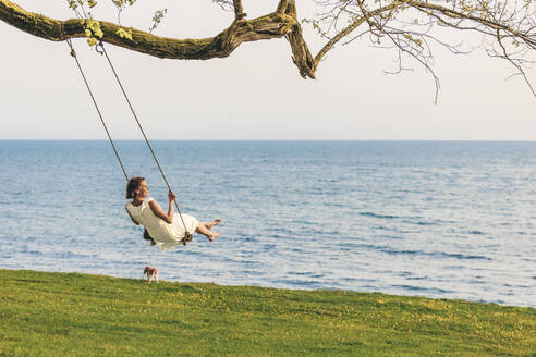 Woman swinging over grass by sea - OMIF00969