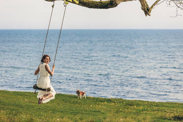 Woman swinging by pet dog standing in front of sea - OMIF00971