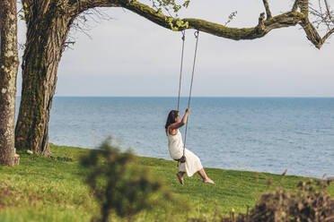 Young woman sitting on swing by sea - OMIF00972