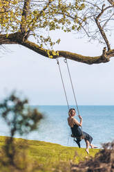 Smiling mature woman swinging in front of sea - OMIF00976