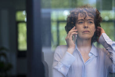 Reife Geschäftsfrau mit Hand im Haar, die mit einem Smartphone spricht, gesehen durch Glas - JOSEF11236