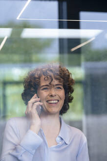 Lächelnde Geschäftsfrau mit lockigem Haar, die mit einem Smartphone spricht, gesehen durch Glas - JOSEF11238