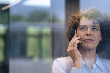 Reife Geschäftsfrau mit lockigem Haar, die mit einem Mobiltelefon spricht, gesehen durch Glas - JOSEF11248