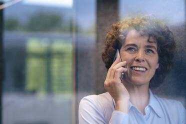 Happy businesswoman talking on mobile phone seen through glass - JOSEF11249