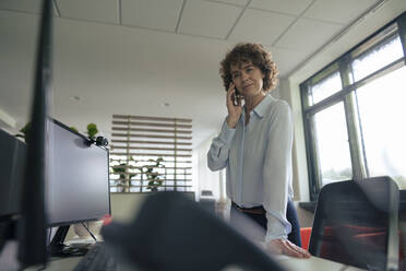 Businesswoman talking on smart phone by desk in office - JOSEF11261