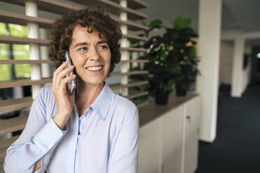 Glückliche Geschäftsfrau mit lockigem Haar, die im Büro mit einem Mobiltelefon spricht - JOSEF11263