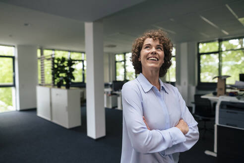 Happy businesswoman standing with arms crossed in office looking up - JOSEF11269