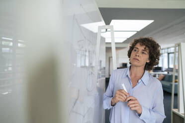 Businesswoman with curly hair looking at whiteboard in office - JOSEF11273