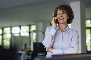 Glückliche Geschäftsfrau mit lockigem Haar und Brille, die im Büro mit einem Mobiltelefon spricht - JOSEF11282