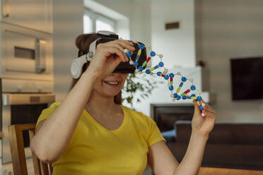 Smiling woman wearing VR goggles and holding DNA model at home - OSF00428