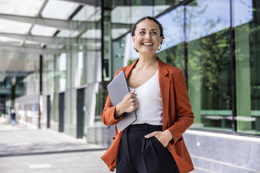 Smiling businesswoman holding laptop standing with hand in pocket - WPEF06199
