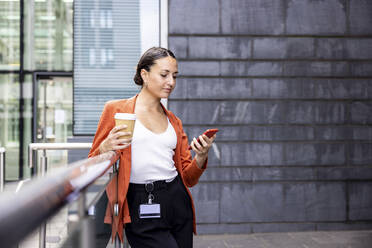 Geschäftsfrau, die einen Einwegbecher hält und auf ein Mobiltelefon am Geländer schaut - WPEF06210