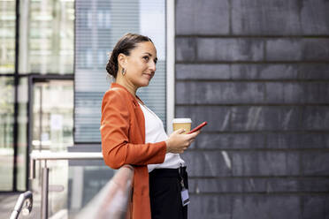 Businesswoman holding disposable cup and smart phone leaning on railing - WPEF06212