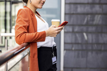 Geschäftsfrau hält Einwegbecher mit Smartphone und lehnt sich an ein Geländer - WPEF06213