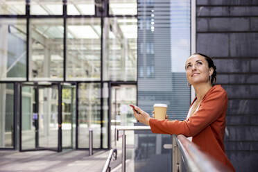 Smiling businesswoman holding disposable cup and smart phone leaning on railing - WPEF06214