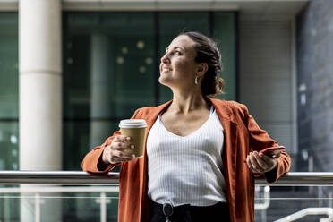 Smiling businesswoman holding smart phone and disposable cup leaning on railing - WPEF06215