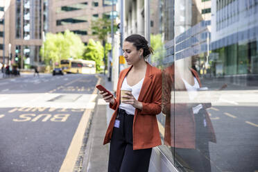 Geschäftsfrau mit Einwegbecher und Smartphone, die sich an eine Glaswand auf dem Gehweg lehnt - WPEF06220