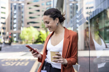 Smiling businesswoman holding disposable cup using smart phone leaning on glass wall - WPEF06221