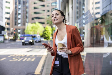 Businesswoman holding disposable cup and smart phone standing by glass wall - WPEF06222