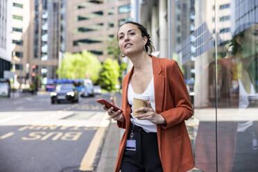 Geschäftsfrau mit Einwegbecher und Smartphone vor einer Glaswand - WPEF06222