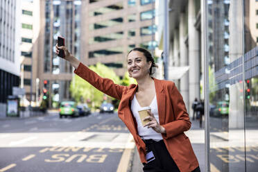 Glückliche Geschäftsfrau mit Einwegbecher und Mobiltelefon in der Hand, die eine Fahrt in der Stadt ankündigt - WPEF06223