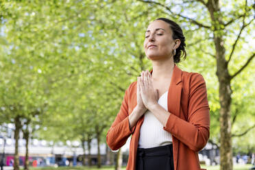 Smiling businesswoman with hands clasped meditating in park - WPEF06228