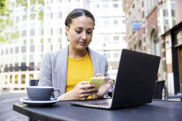 Businesswoman using smart phone sitting with laptop at sidewalk cafe - WPEF06251